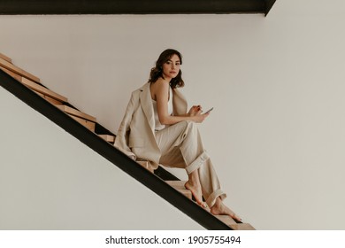 Young Brunette Curly Woman In Beige Suit Sits On Wooden Stairs. Pretty Tanned Lady In Linen Pants And Jacket Holds Phone.