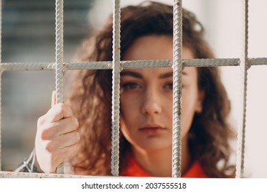 Young Brunette Curly Woman In Orange Suit Behind Jail Bars. Female In Colorful Overalls Portrait