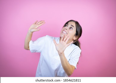 Young brunette doctor girl wearing nurse or surgeon uniform over isolated pink background scared with her arms up like something falling from above