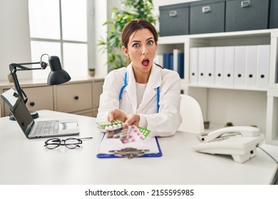 Young Brunette Doctor Woman Holding Pills In Shock Face, Looking Skeptical And Sarcastic, Surprised With Open Mouth 