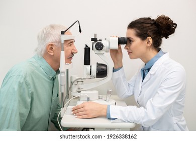 Joven morena oftalmóloga femenina sentada por equipo médico en clínicas mientras se examina la vista de paciente masculino de mayor edad frente a ella