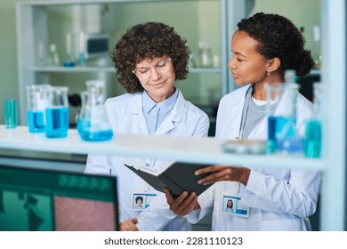 Young brunette female researcher in lab coat and eyeglasses looking at notes in notebook held by African American colleague