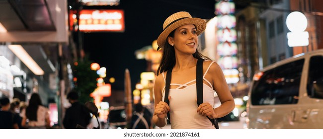 Young Brunette Female Solo Traveler Walking On City Street In Evening Night In Summer Clothes. Happy And Excited 20s Hispanic Girl Backpacker On Holiday Vacation Alone. Solo Traveling Woman Concept