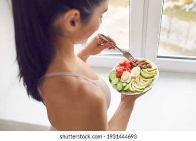 Young Brunette Fitness Woman Athete In Sportswear Standing And Eating Healthy Meal With Eggs, Nuts And Vegetables Before Or After Workout At Home, Top View. Active Healthy Lifestyle, Clean Eating