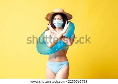 young brunette girl in a blue swimsuit with a swimming inflatable ring puts on a medical mask on a yellow background and shows a stop gesture
