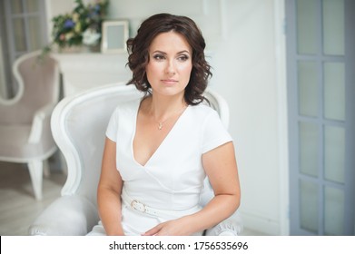 young brunette girl with curly hair in a white dress sits in a beautiful armchair, looks out the window. Beautiful brown eyes. Mother's Day, Women's Day. Birthday. Close-up