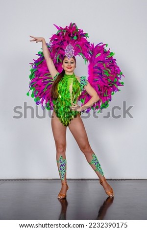 Young brunette girl dancer in a carnival costume of green and pink feathers poses on a white background
