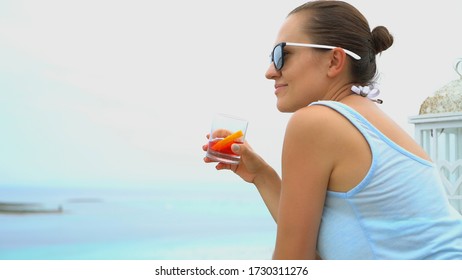 Young brunette girl drinking red cocktail from a glass on outdoor terrace by the sea