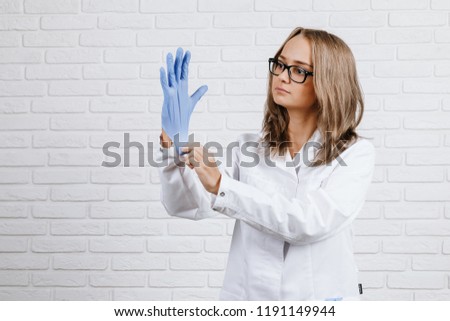 Young brunette girl with flowing hair in black glasses in a white robe puts on a medical glove on her right hand on a white background