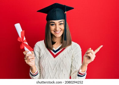 Young brunette girl holding graduate degree diploma smiling happy pointing with hand and finger to the side 