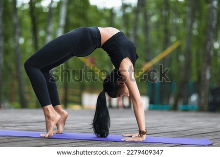 A young brunette girl leading a healthy lifestyle and practicing yoga, performs the Setu Bandha Sarvangasana exercise, bridge pose, trains in black sportswear on a mat in the park