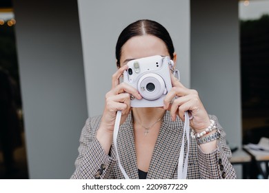 A Young Brunette Girl Poses With A Polaroid Camera Against A Gray Wall Background. High Quality Photo