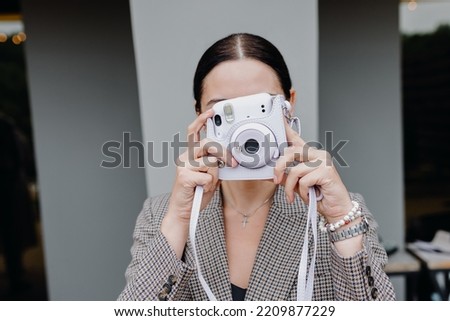 A young brunette girl poses with a polaroid camera against a gray wall background. High quality photo