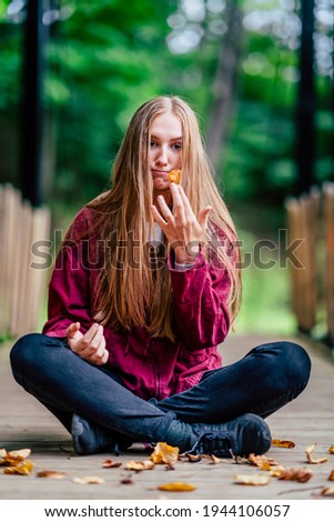 Young Brunette Girl Sits on Ground, Holds Fallen Leaf, Makes Funny Face Expression. Purple Sweater and Dark Blue Jeans