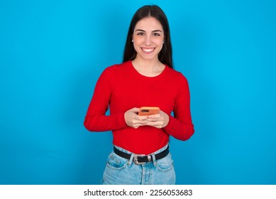 young brunette girl wearing red T-shirt against blue wall enjoys distant communication, uses mobile phone, surfs fast unlimited internet, has pleasant smile, makes shopping online, - Shutterstock ID 2256053683