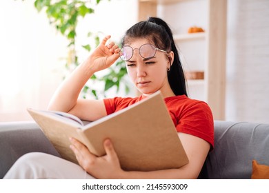 A Young Brunette With Glasses Is Reading A Book, Having Poor Eyesight At Home. A Student Experiencing Vision Problems Or Eye Fatigue Is Reading A Book.