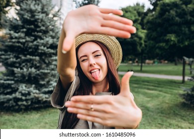 Young Brunette Hipster Woman Taking A Selfie, Showing Tongue, Funny Face Posing Outdoors. Closeup Portrait Of A Girl In A Stylish Summer Hat In A Park.
