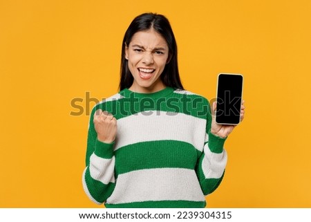 Young brunette latin woman wear casual green knitted sweater hold in hand use mobile cell phone with blank screen workspace area do winer gesture isolated on plain yellow background studio portrait