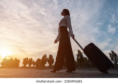 Young Brunette With Long Hair In Businesswear On High Heels Walks Pulling Black Suitcase. Woman Goes On Road Against Cloudy Sky Low Angle Shot, Sinlight