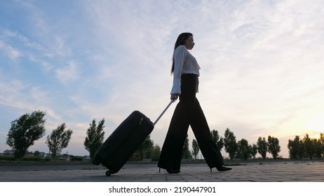 Young Brunette With Long Hair In Businesswear On High Heels Walks Pulling Black Suitcase. Woman Goes On Road Against Cloudy Sky Low Angle Shot