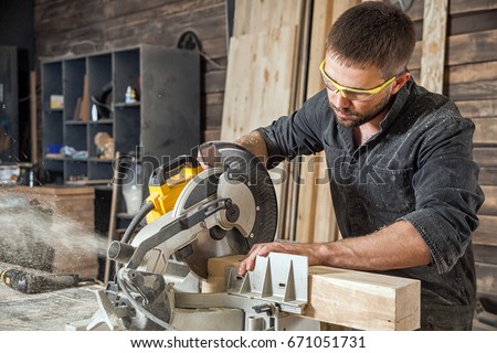Young brunette man in black overalls by profession carpenter builder saws with a circular saw a wooden board on a wooden table in the workshop
