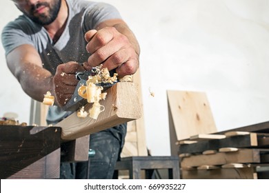 A young brunette man builder in green glasses and a gray T-shirt handles a wooden bar with a black jack plane in the workshop, in the background a lot of wooden boards
