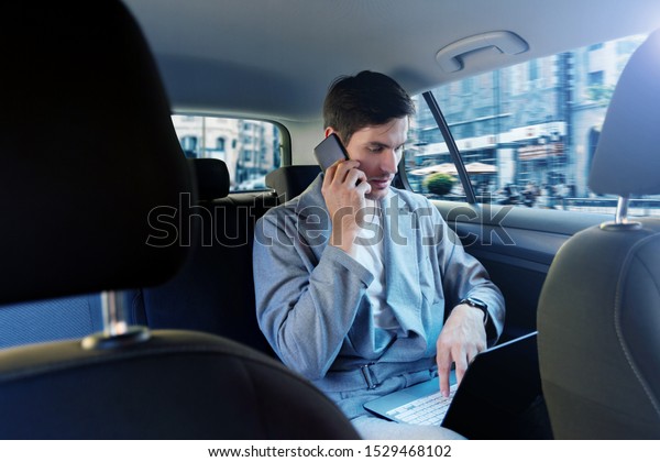 Young brunette man in grey suit is riding in taxi\
in back seat of automobile. Businessman is talking on smartphone,\
working on laptop on way to meeting. Concept of fast rhythm of\
modern city.