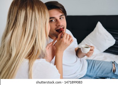 Young Brunette Man Holding Cup Of Coffee In Hand Dreamily Eating Strawberry And Looking At Blond Woman Feeding Him. Couple In White T-shirts Having Breakfast In Bed In Modern Hotel