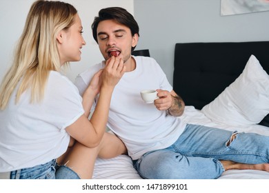 Young Brunette Man Holding Cup Of Coffee In Hand Dreamily Eating Strawberry While Pretty Blond Woman Feeding Him. Beautiful Couple In White T-shirts Having Delicious Breakfast In Bed In Modern Hotel