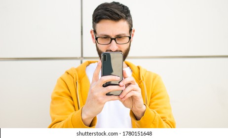 Young Brunette Man Holding Smartphone In Hands In Front Of Him.Close-up Image Of Mobile Phone.Focused Male In Eyeglasses,yellow Hoodie Using Cellphone,making Video Call,selfie,taking Picture,shooting