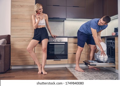 Young Brunette Man Putting Plates In Dishwasher While Looking At Blond Woman Eating Red Apple