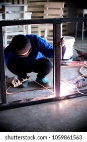 A young brunette man welder in a black T-shirt, in a welding mask and welder leathers, welds a metal welding machine in the workshop, sparks fly in the sides