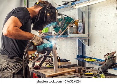A young brunette man welder in a black T-shirt, in a welding mask and welder leathers, welds a metal welding machine in the workshop, sparks fly in the sides