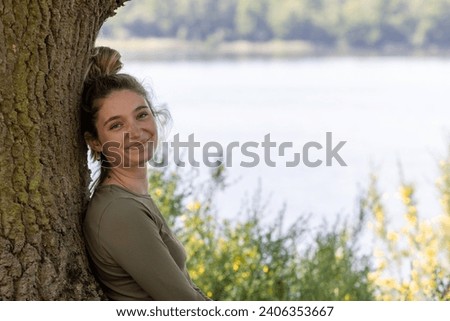 A young brunette millennial woman finds serenity in nature's embrace. She sits by a forest lake, leaning against a tree, enjoying the tranquility of the lush green surroundings on a summer day