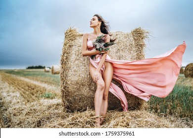 Young brunette in a pink satin dress, without linen standing near hay bales in windy weather. A woman holds a bouquet of wildflowers. - Shutterstock ID 1759375961