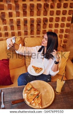 Young brunette sitting indoors with pizza and smartphone in hand.