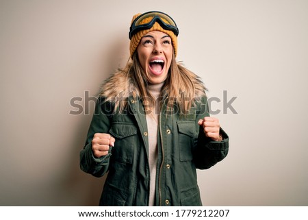 Young brunette skier woman wearing snow clothes and ski goggles over white background angry and mad screaming frustrated and furious, shouting with anger. Rage and aggressive concept.
