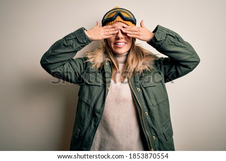 Young brunette skier woman wearing snow clothes and ski goggles over white background covering eyes with hands smiling cheerful and funny. Blind concept.