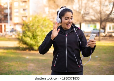 Young Brunette Sporty Girl Using Headphones Walking At The Park And Saying Hi Very Happy