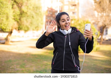 Young Brunette Sporty Girl Wearing Headphones Walking At The Park Using Smartphone And Saying Hi Very Happy