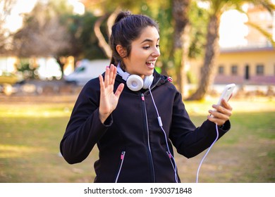 Young Brunette Sporty Girl Wearing Headphones Walking At The Park Using Smartphone And Saying Hi Very Happy