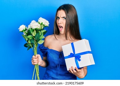 Young Brunette Teenager Holding Anniversary Present And Bouquet Of Flowers In Shock Face, Looking Skeptical And Sarcastic, Surprised With Open Mouth 