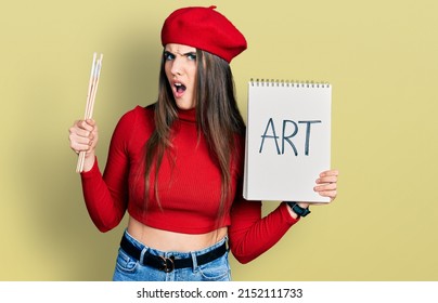 Young Brunette Teenager Holding Art Notebook And Paintbrushes In Shock Face, Looking Skeptical And Sarcastic, Surprised With Open Mouth 