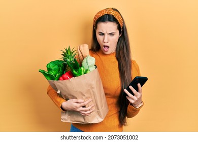 Young Brunette Teenager Holding Bag Of Groceries Using Smartphone In Shock Face, Looking Skeptical And Sarcastic, Surprised With Open Mouth 