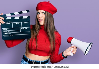 Young Brunette Teenager Holding Video Film Clapboard And Megaphone Clueless And Confused Expression. Doubt Concept. 