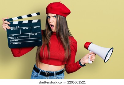 Young Brunette Teenager Holding Video Film Clapboard And Megaphone In Shock Face, Looking Skeptical And Sarcastic, Surprised With Open Mouth 