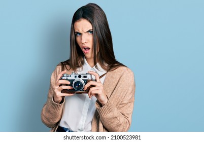 Young Brunette Teenager Holding Vintage Camera In Shock Face, Looking Skeptical And Sarcastic, Surprised With Open Mouth 