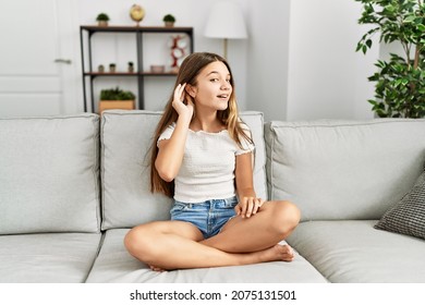 Young brunette teenager sitting on the sofa at home smiling with hand over ear listening an hearing to rumor or gossip. deafness concept.  - Shutterstock ID 2075131501
