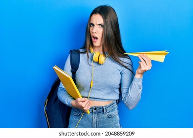 Young Brunette Teenager Student Holding Books And Paper Plane In Shock Face, Looking Skeptical And Sarcastic, Surprised With Open Mouth 