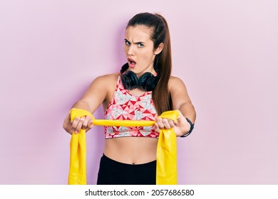 Young Brunette Teenager Training Arm Resistance With Elastic Arm Bands In Shock Face, Looking Skeptical And Sarcastic, Surprised With Open Mouth 